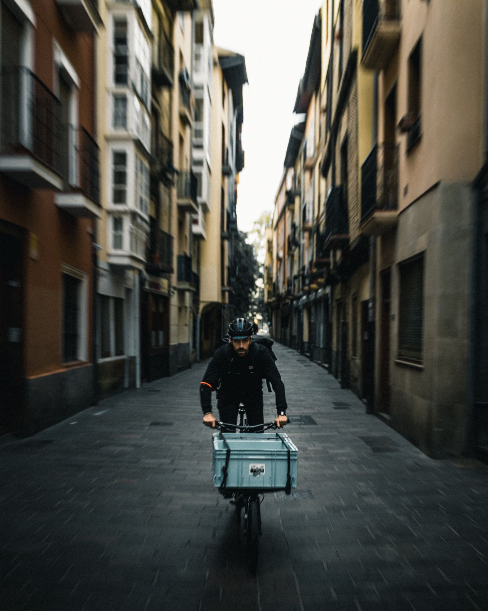 Un repartidor de frente por una calle estrecha, con la caja en la bici